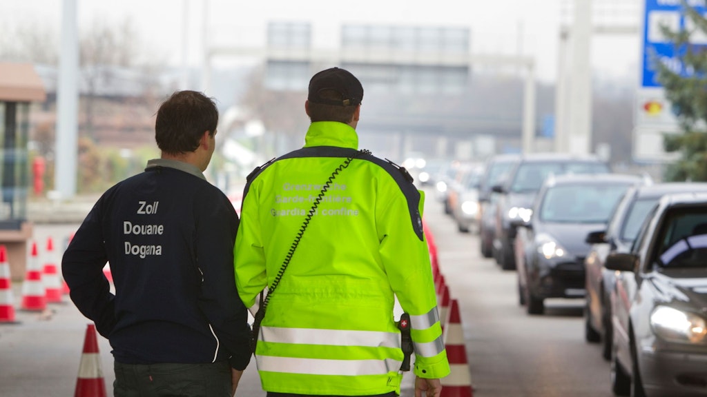 Deux hommes se tiennent au bord d'une route et observent les voitures qui passent. Les voitures forment une longue file, elles sont éclairées et on ne les distingue pas clairement. L'un des deux hommes porte une veste bleu foncé avec l'inscription "Zoll, Douane, Dogana" et a les cheveux courts et foncés. L'autre homme porte une veste jaune fluorescente avec l'inscription "Grenzwache, Garde-frontière, Guardia di confine" et une casquette Baselball noire. On ne voit que le dos et l'arrière de la tête des deux hommes.