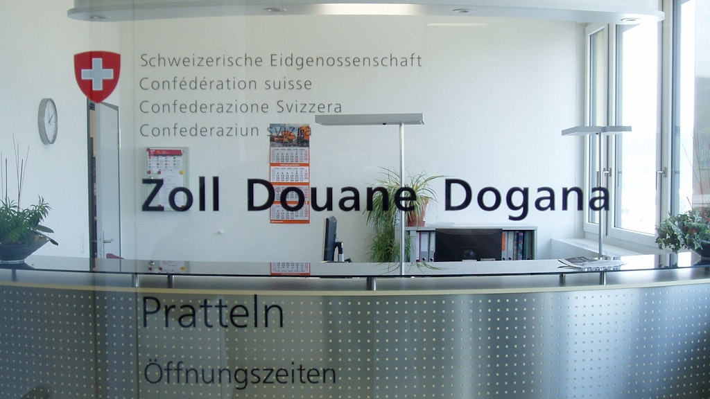 Porte vitrée dans un immeuble de bureaux avec le logo Schweizerische Eidgenossenschaft, Confédération suisse, Confederazione Svizzera, Confederaziun svizra et la croix suisse. En dessous sont imprimés les mots Zoll, Douane, Dogana, Pratteln et Öffnungszeiten. Derrière la porte vitrée, on voit un guichet avec des brochures d'information et des plantes. Derrière le guichet, on peut voir deux écrans et un caisson avec plusieurs classeurs. Une horloge, un calendrier et un tableau magnétique avec plusieurs feuilles d'information sont accrochés au mur.
