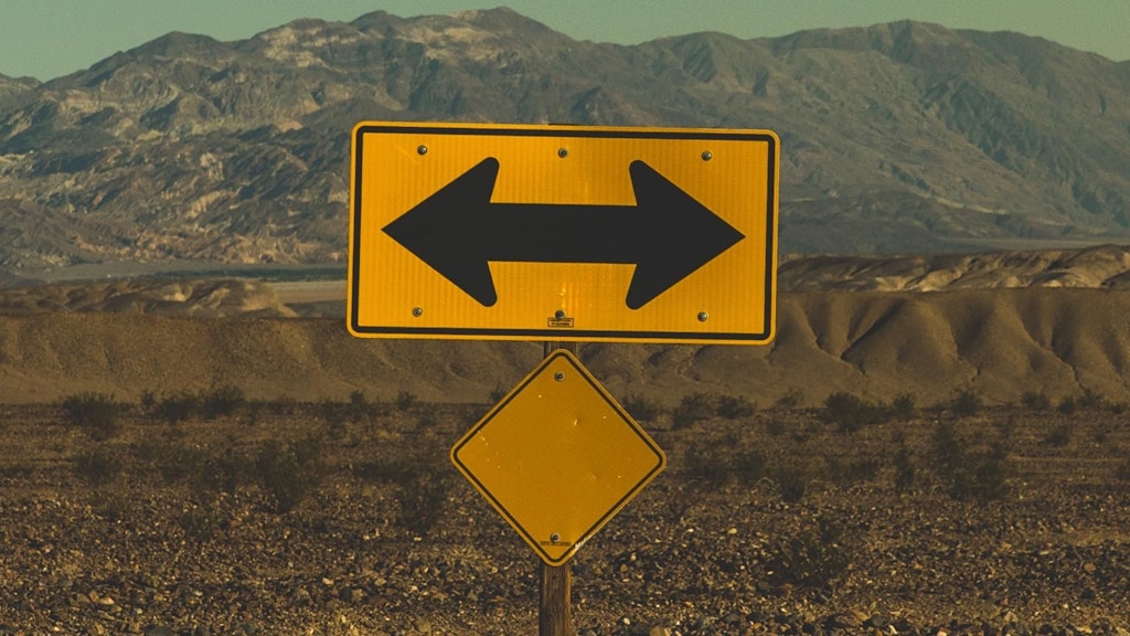 Gelbes Schild mit schwarzen Doppelpfeil. Im Hintergrund ist eine bergige, Karge Landschaft zu sehen