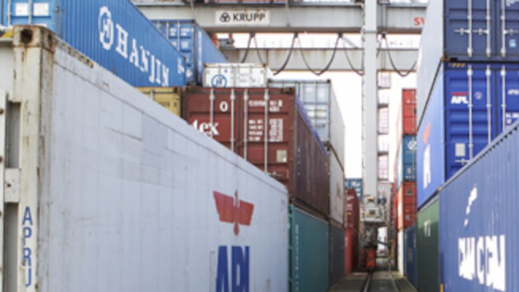 Numerosi container di diversi colori sono impilati accanto a un binario ferroviario. Sopra i container è visibile una gru per container.