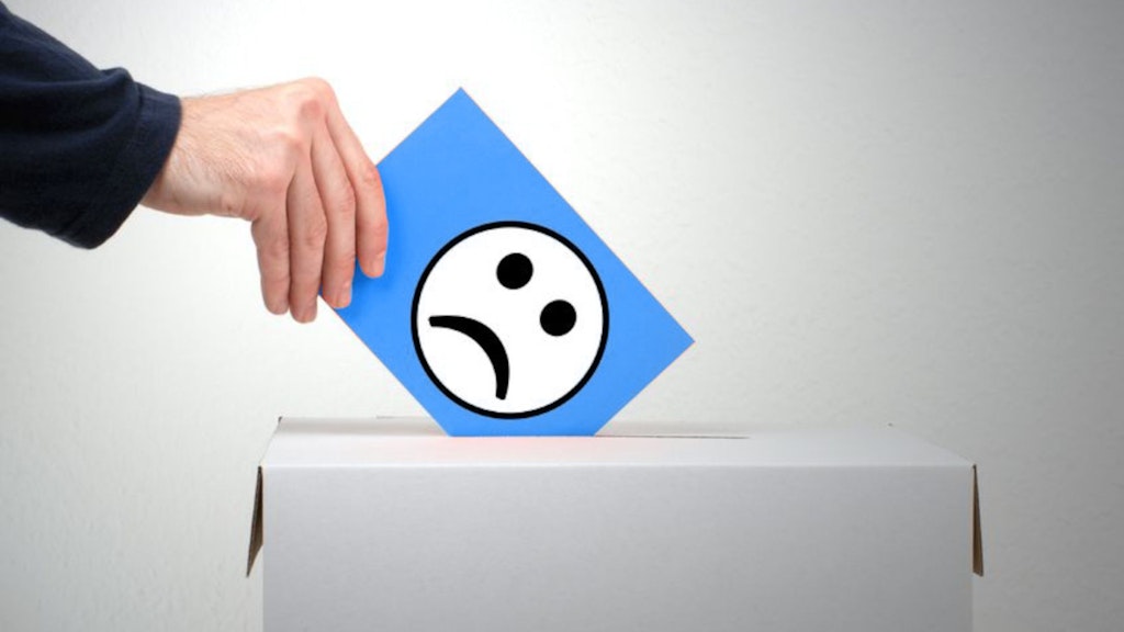 In questa immagine, qualcuno sta gettando un biglietto blu con una faccina triste in una scatola grigia. Questa scatola è chiusa e ha una fessura come una cassetta delle lettere. La scatola si trova di fronte a una parete grigia. Della persona si vede solo la mano destra e parte del braccio, coperto da una manica blu scuro.