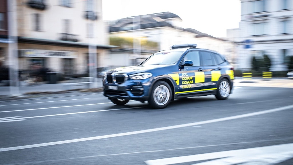 Ein Zollfahrzeug fährt schnell auf einer Straße in der Stadt. Es ist ein dunkelblauer SUV mit gelben Markierungen und der Aufschrift "Zoll" auf der Seite.