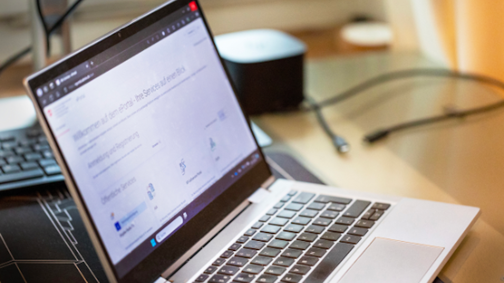 Un ordinateur portable ouvert affiche la page d'accueil de la plateforme fiscale « Taxas » avec différents services numériques. L'écran est légèrement flou, mais la structure de la plateforme est visible.