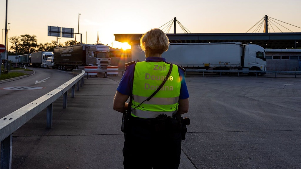 Une personne en gilet de douane se tient au lever du soleil sur un parking, tandis que des camions sont garés