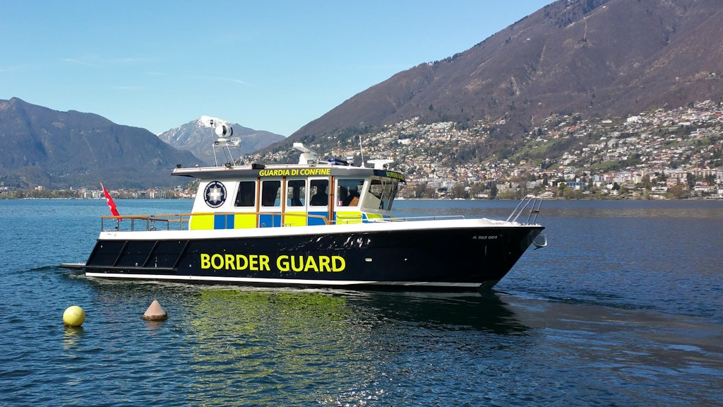 Un bateau de la garde frontière navigue sur un lac avec des montagnes en arrière-plan. 