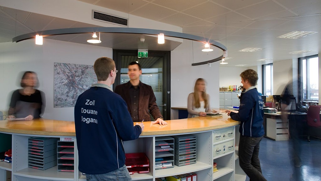 Two customs officers stand at a reception desk and speak with visitors. Other people and office desks are visible in the background.