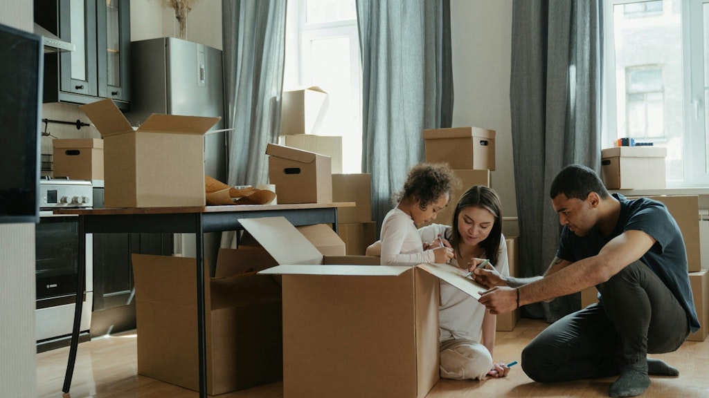 A family sits in a room full of moving boxes. A child and two adults are busy unpacking and organizing.