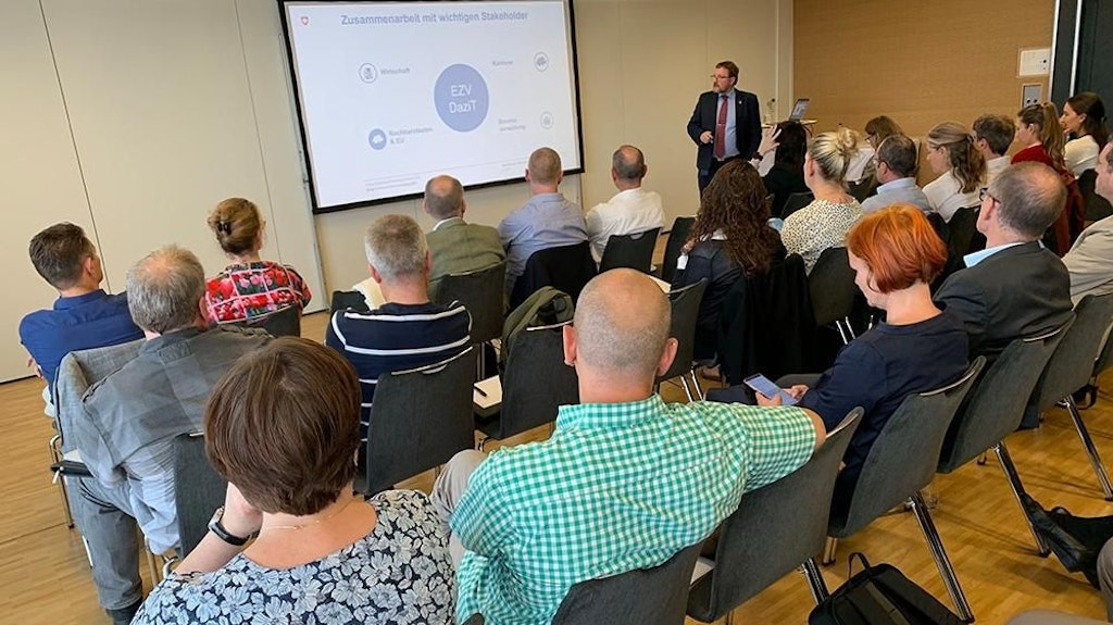 Un groupe de personnes est assis dans une salle de conférence et écoute une présentation. Un diaporama sur le programme DaziT de l'Office fédéral de la douane et de la sécurité des frontières est visible à l'écran.