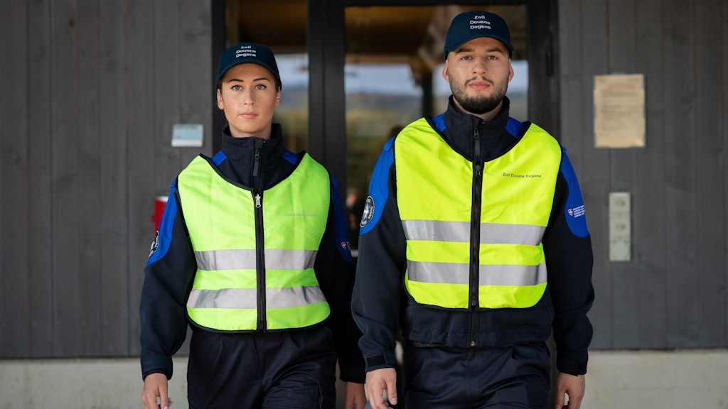 Deux spécialistes en douane et sécurité des frontières en uniforme se tiennent devant un bâtiment. Ils portent des gilets réfléchissants et des casquettes avec l'inscription "Zoll Douane Dogana".