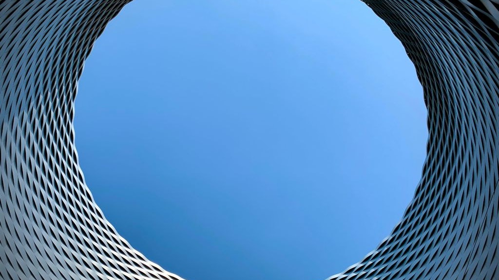 Blick durch eine runde Öffnung einer modernen Metallfassade in den blauen Himmel. Die Struktur bildet ein dynamisches, spiralförmiges Muster.  Caption-de: Runde Öffnung in einer modernen Metallfassade mit Sicht in den klaren blauen Himmel. Die architektonische Struktur wirkt abstrakt und futuristisch.  