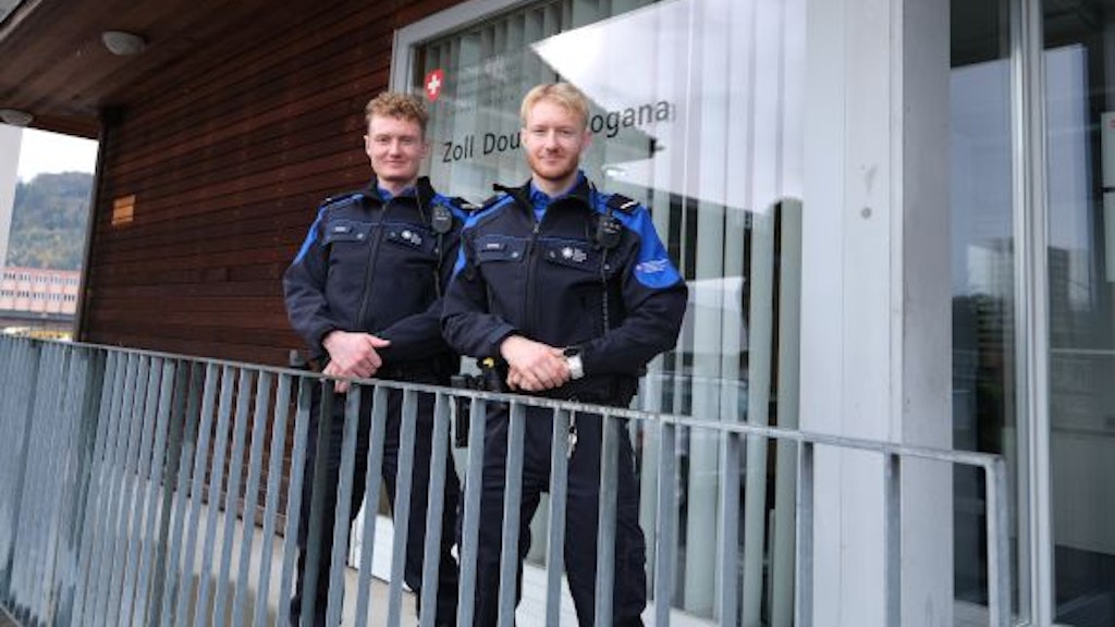 Deux douaniers se tiennent devant un bâtiment de bureaux avec l'inscription "Zoll Douane Dogana". Ils portent des uniformes et sourient à la caméra.
