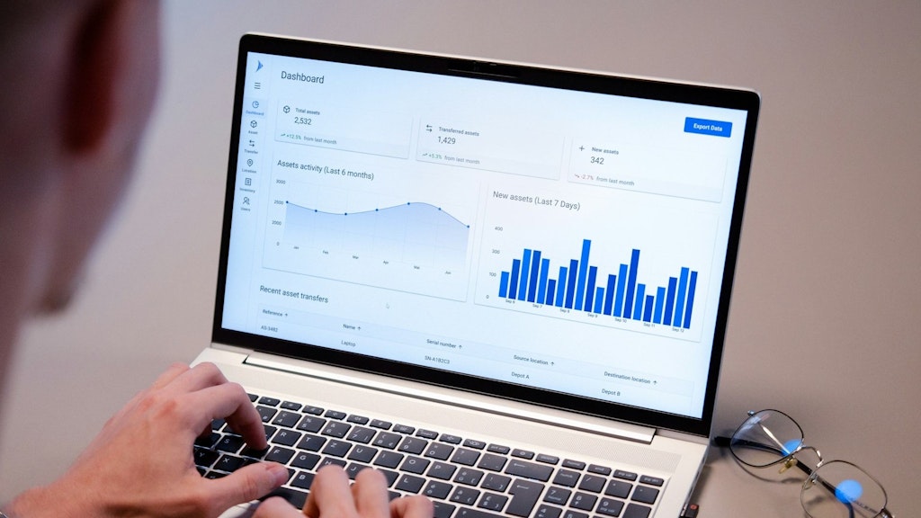 Ein Laptop zeigt ein Dashboard mit Diagrammen und Statistiken. Eine Person tippt auf der Tastatur, während eine Brille daneben liegt.