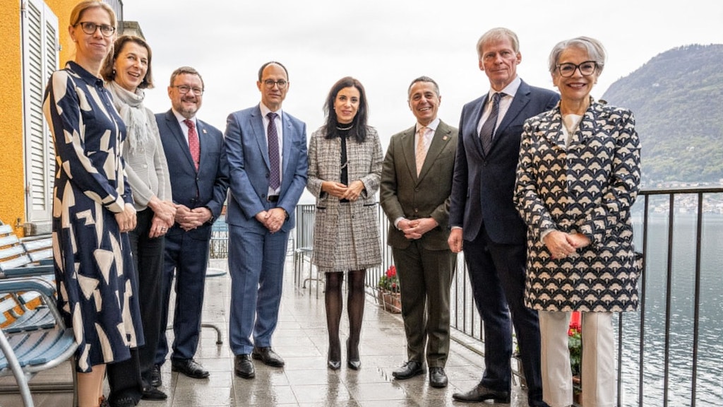 Plusieurs personnes se tiennent côte à côte lors d’un vernissage sur une terrasse avec vue sur un lac et regardent l’objectif en souriant. En arrière-plan, on aperçoit l’eau et les montagnes.