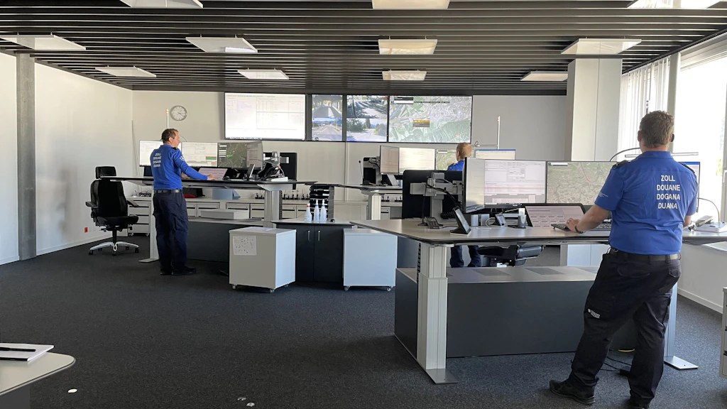 Plusieurs collaborateurs de la douane en uniforme bleu travaillent dans la centrale d'engagement Est, dans un grand local moderne équipé de nombreux écrans. De grands moniteurs muraux affichent des cartes et des images vidéo de la région frontalière, tandis que la lumière du jour entre par une large baie vitrée.  