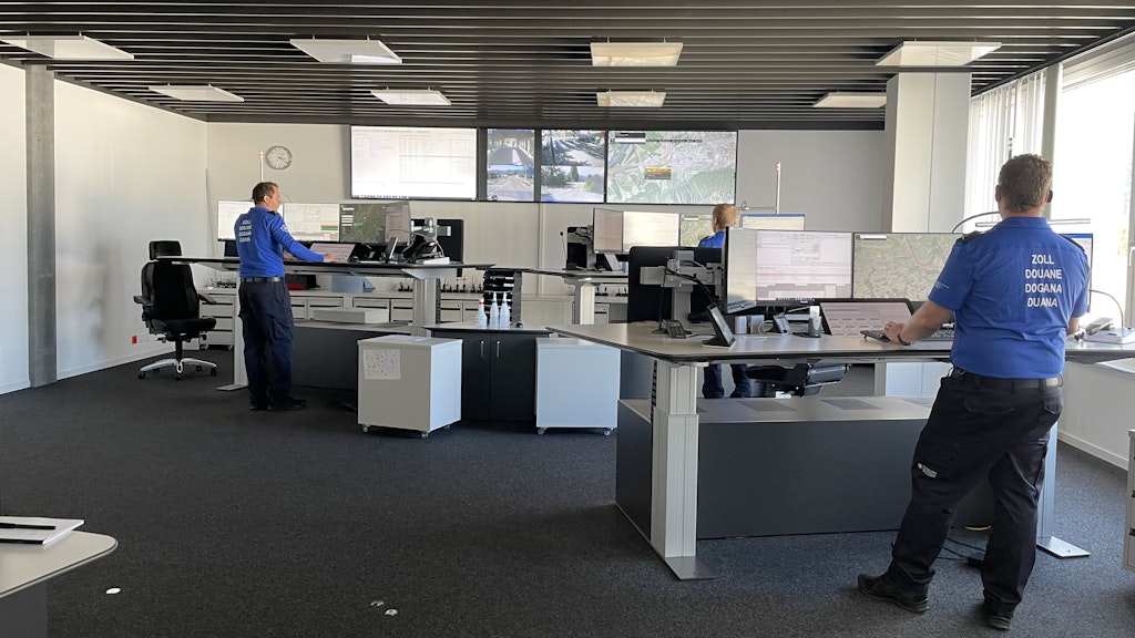 Mehrere Zollmitarbeitende in blauen Uniformen arbeiten in einer moder-nen Einsatzzentrale Ost an höhenverstellbaren Pulten mit zahlreichen Monitoren. Auf grossen Bildschirmen an der Front sind Karten und Kame-rabilder des Grenzgebiets zu sehen. Durch die Fenster fällt Tageslicht in den grosszügigen Raum.  