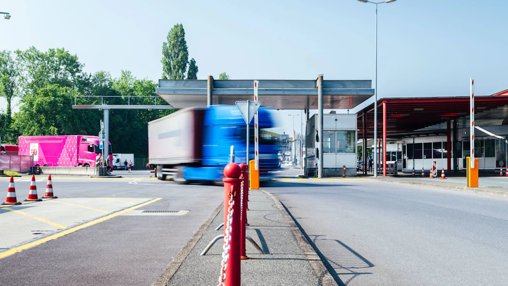  Un camion bleu traverse un poste frontière moderne. En arrière-plan, un camion rose et plusieurs guérites de contrôle sont visibles, ainsi que des cônes de signalisation. La scène illustre le trafic de marchandises à une frontière.  