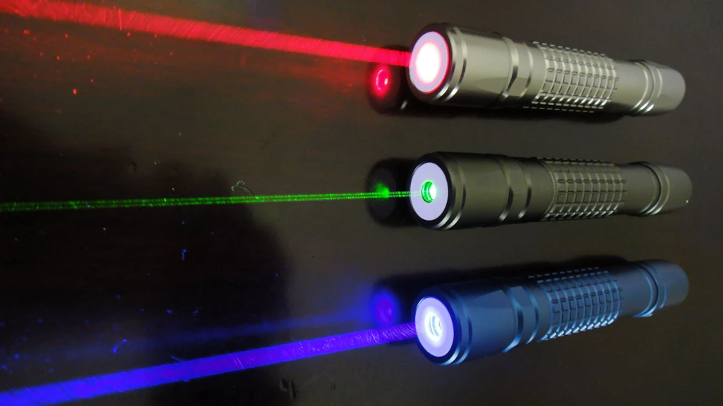 Three switched-on laser pointers are lying side by side on a dark surface. The laser beams are clearly visible in red, green, and blue. Each pointer emits a distinct light beam in its respective color.