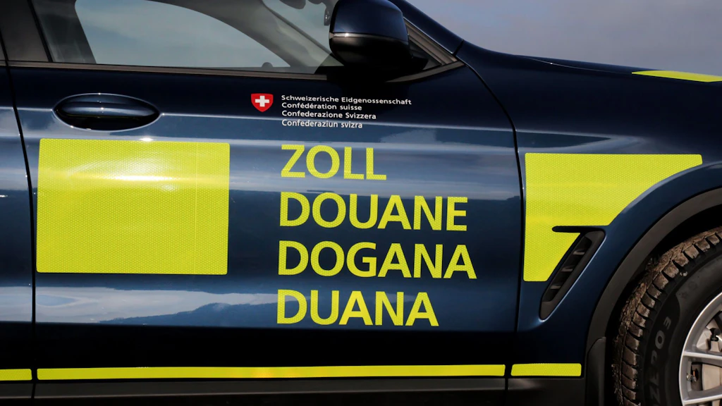 n véhicule moderne de la douane suisse avec des inscriptions jaunes dans les quatre langues nationales est garé à l'extérieur. Sur la porte, on peut lire clairement "Zoll", "Douane", "Dogana" et "Duana". Le véhicule porte l'emblème de la Confédération suisse.