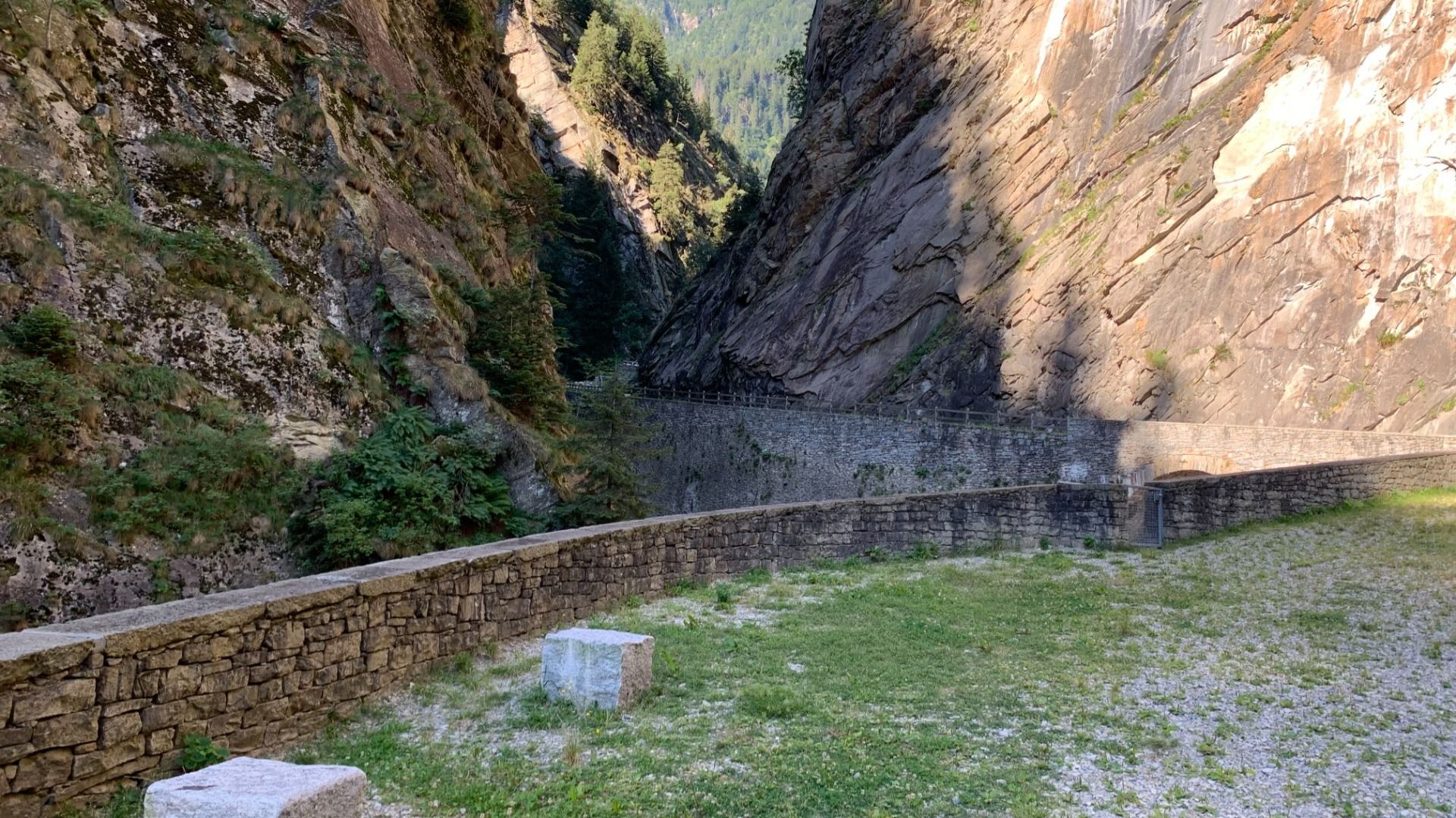 Ein steiniger Bergpass mit hohen Felswänden und einer alten Steinmauer. Die Umgebung ist von grüner Vegetation umgeben.  