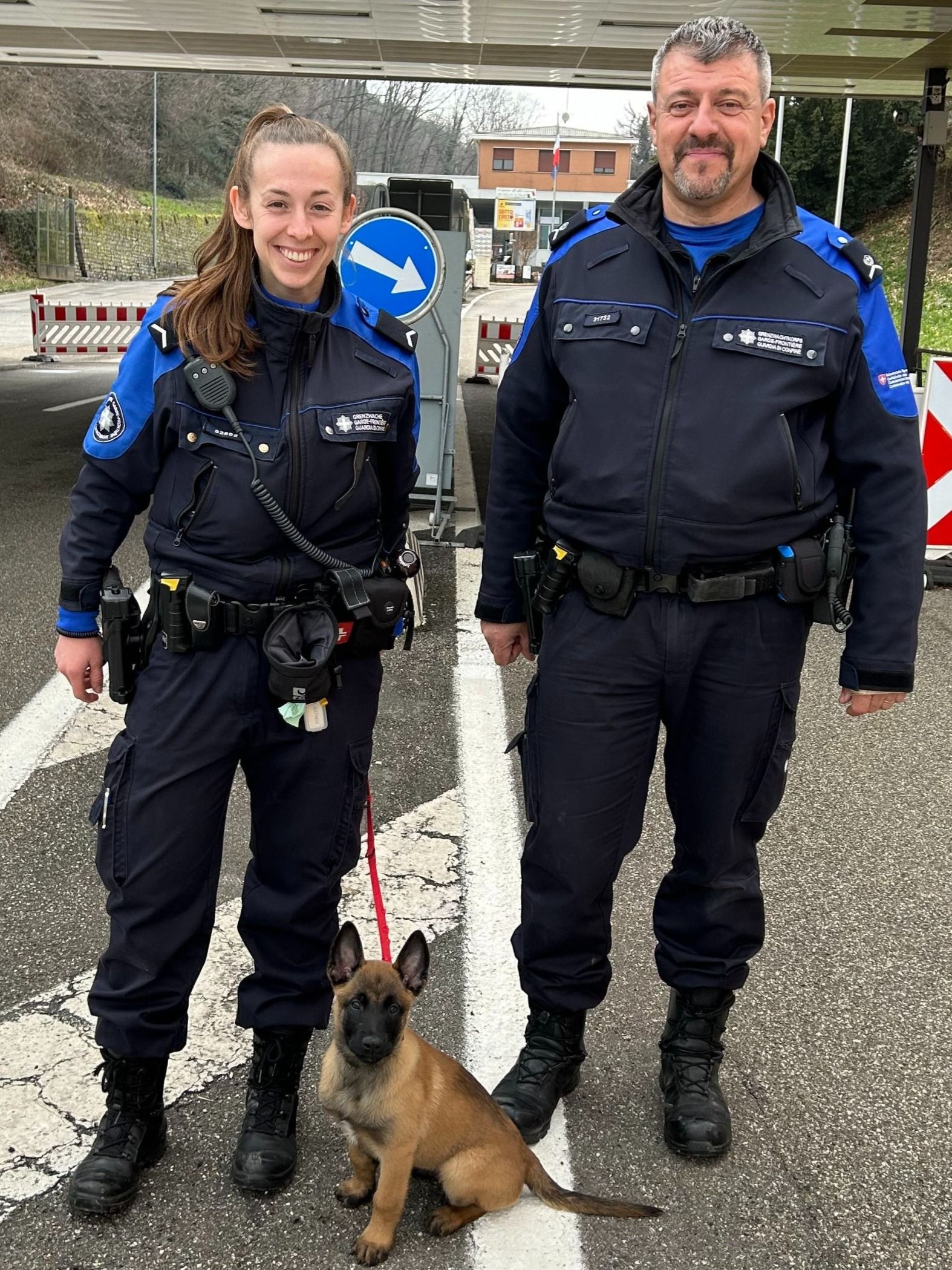 Zwei Personen in Uniform stehen mit einem Hund an einem Grenzübergang.  