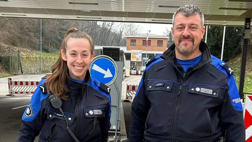 Zwei Personen in Uniform stehen mit einem Hund an einem Grenzübergang.  