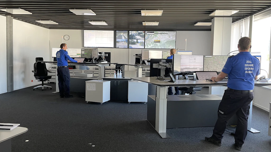 Mehrere Zollmitarbeitende in blauen Uniformen arbeiten in einer modernen Einsatzzentrale Ost an höhenverstellbaren Pulten mit zahlreichen Monitoren. Auf grossen Bildschirmen an der Front sind Karten und Kamerabilder des Grenzgebiets zu sehen. Durch die Fenster fällt Tageslicht in den grosszügigen Raum.  