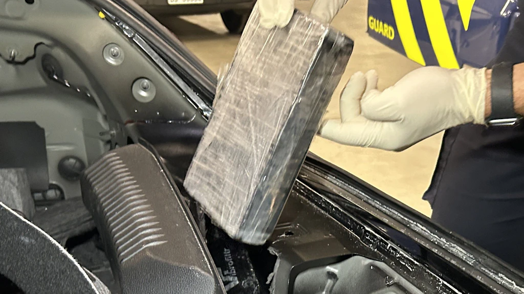 Eine Person mit Handschuhen hält ein in Plastik verpacktes Paket, das aus einem versteckten Fach im Auto entnommen wird. Das Bild zeigt eine Kontrolle durch die Zollbehörde. Im Hintergrund ist ein Fahrzeug mit gelben Streifen sichtbar.