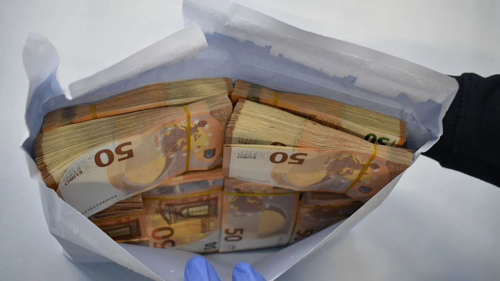 Ein geöffneter Umschlag mit mehreren Bündeln von 50-Euro-Scheinen, gehalten von einer Person mit blauen Handschuhen. Die Geldscheine sind ordentlich gestapelt und mit Gummibändern fixiert. Der Hintergrund ist neutral und hell.