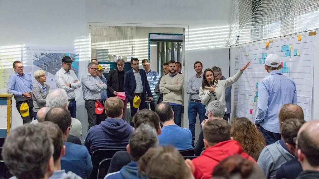 Eine Gruppe von Menschen versammelt sich in einem Raum für ein PI Planning Meeting. Eine Frau spricht und zeigt auf ein grosses Diagramm an der Wand. Die Teilnehmer hören aufmerksam zu.