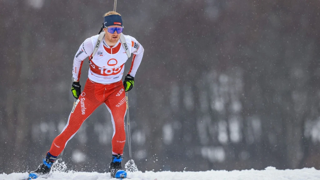 Ein männlicher Biathlet in rotem Rennanzug und Startnummer läuft auf Skiern durch den Schnee. Er trägt eine Schutzbrille und ein Stirnband, während er sich auf den Wettkampf konzentriert. Im Hintergrund sind verschwommene Bäume und fallender Schnee zu sehen.  