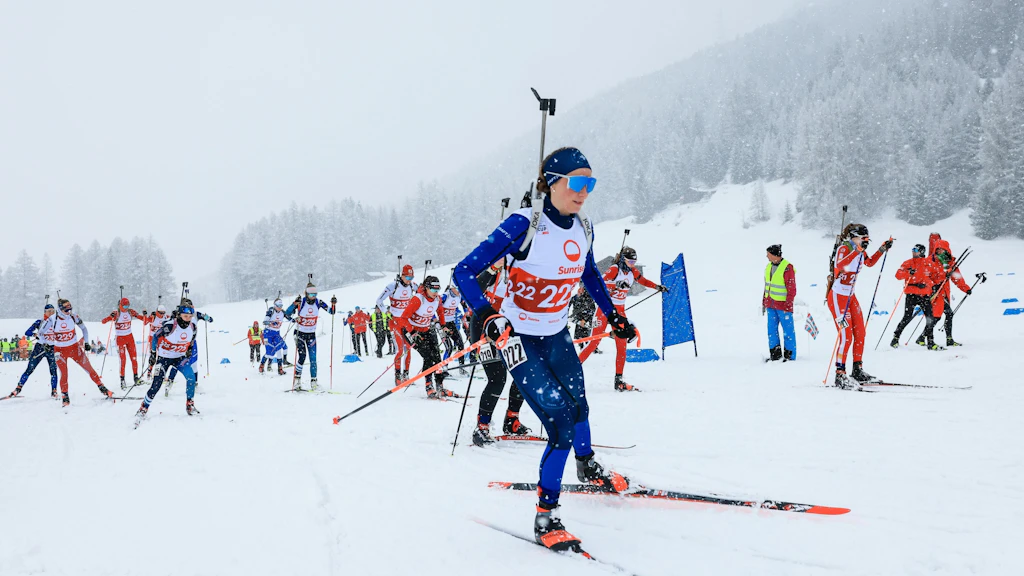 Mehrere Biathletinnen und Biathleten starten bei starkem Schneefall in ein Rennen in Ulrichen. Im Vordergrund laeuft eine Athletin in blauem Rennanzug mit Gewehr auf dem Ruecken, im Hintergrund sind weitere Laeuferinnen, Betreuende und verschneite Baeume zu sehen.