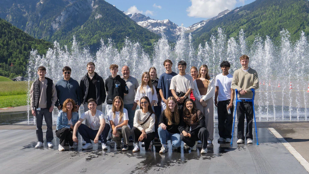 Eine Gruppe von Menschen posiert vor einem sprudelnden Brunnen mit Bergen im Hintergrund. Sie stehen auf einer nassen Fläche und lächeln in die Kamera.  