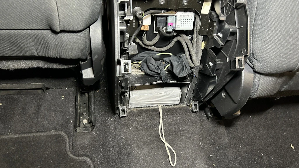 In einem Auto wurde ein Versteck unter einem Sitz entdeckt, in dem ein in Plastik verpacktes Paket mit Kokain sichtbar ist. Das Fach ist geöffnet und zeigt Kabel sowie das Paket mit einer herausragenden Schnur.