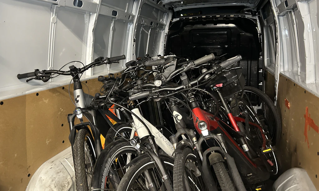 Mehrere Fahrräder sind dicht nebeneinander im Laderaum eines Lieferwagens verstaut. Die Fahrräder stehen aufrecht und nehmen den gesamten hinteren Bereich des Fahrzeugs ein. Die Umgebung wirkt wie ein typischer Transporter-Innenraum.  
