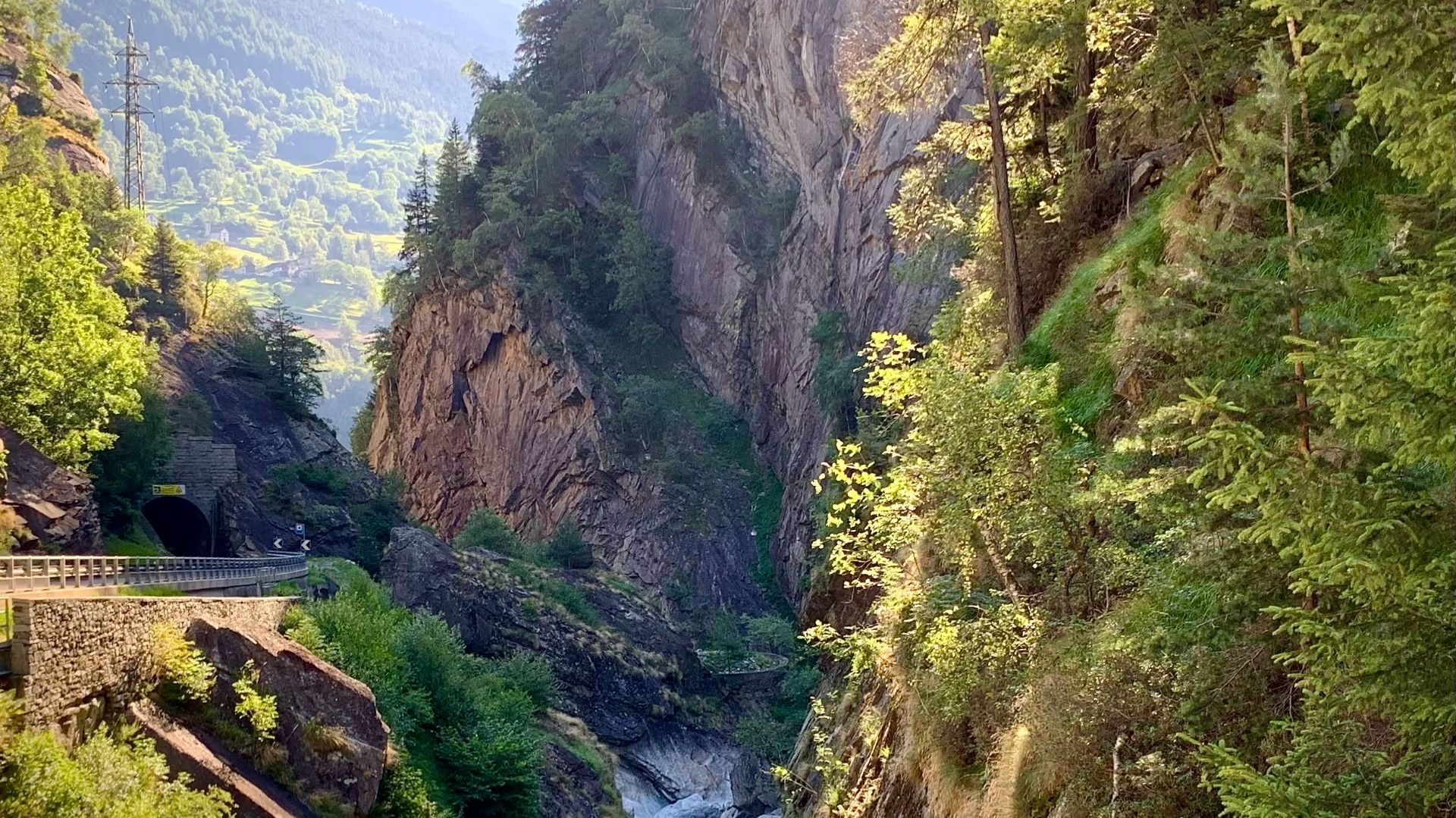 Eine malerische Bergschlucht mit einem Tunnel, umgeben von üppigem Grün und steilen Felswänden. Im Hintergrund sind bewaldete Hügel zu sehen.  