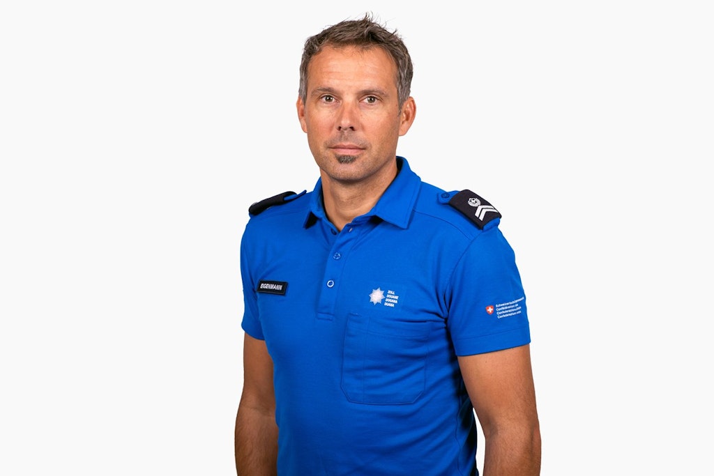 Ein Mann in einer blauen Uniform mit Abzeichen steht vor einem weissen Hintergrund. Er trägt ein Namensschild mit der Aufschrift "Eigenmann". 