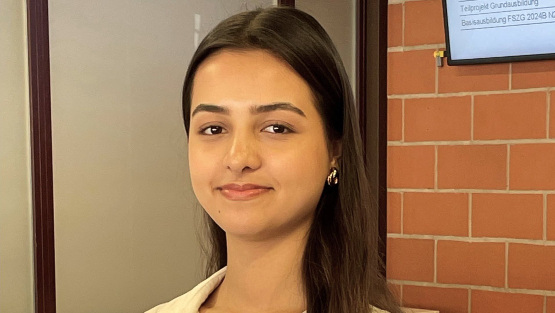 Eine junge Frau mit langen Haaren lächelt in die Kamera. Sie steht vor einer Wand mit einem Bildschirm. 