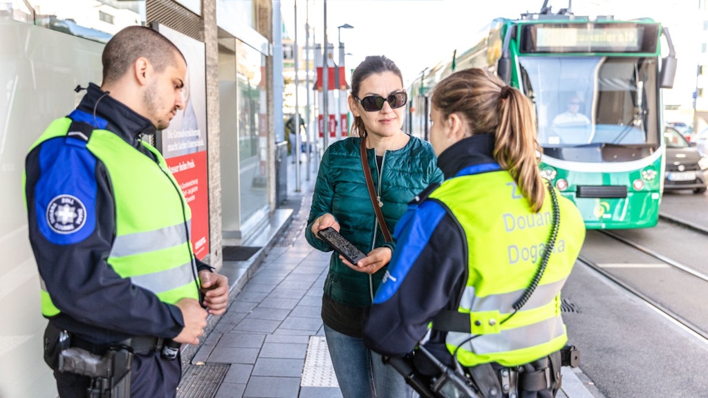 Eine Passantin mit dunkelgrüner Jacke, schwarzer Sonnebrille und dunklen Haaren unterhält sich mit zwei Mitarbeitenden des Zolls. Sie zeigt Ihnen ihr schwarzes Smartphone. Die beiden Mitarbeitenden des Zolls sind eine Frau und ein Mann. Die Frau wendet der Kamera den Rücken zu und trägt ihre langen blonden Haare zusammengebunden. Der Mann steht seitlich zur Kamera, hat kurze dunkle Haare und einen Drei-Tage-Bart. Die Mitarbeitenden tragen eine blaue Uniform, eine gelbe Leuchtweste mit der Aufschrift Zoll, Douane, Dogana und sind bewaffnet. Im Hintergrund steht eine grüne Strassenbahn.