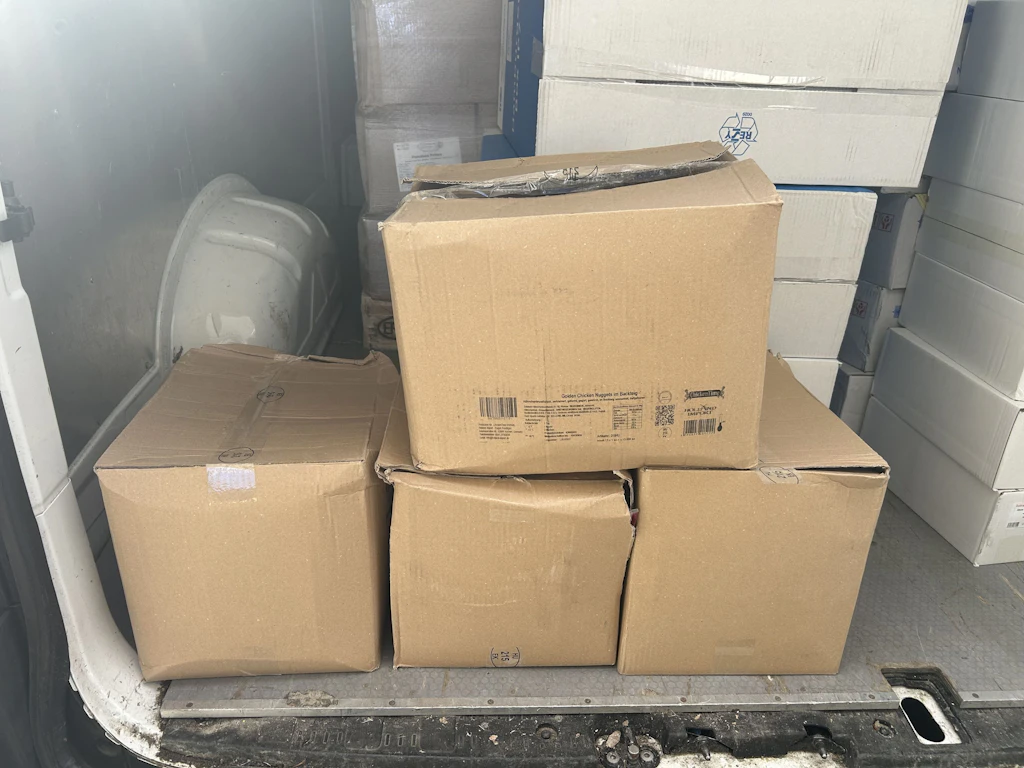 Vier braune Kartons stehen im Laderaum eines Lieferwagens. Im Hintergrund sind weitere weisse Kartons gestapelt. Die Kartons wirken teilweise eingedrückt und sind für den Transport vorbereitet.  