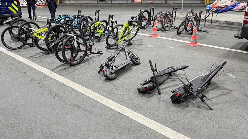 Mehrere Fahrräder und E-Scooter liegen auf einem Zollplatz, offenbar von den Behörden sichergestellt. Im Hintergrund stehen zwei Personen in Uniform und ein Zollfahrzeug. Die Szene ist mit Verkehrskegeln abgesperrt.