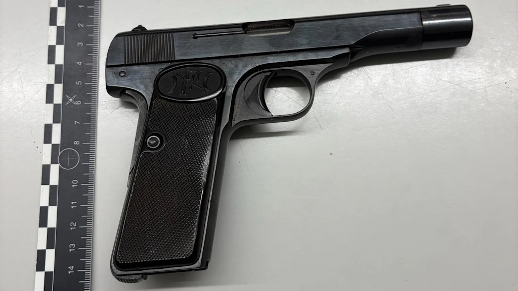 Eine schwarze Pistole liegt auf einer hellen Oberfläche neben einem Massstab. Die Pistole ist seitlich abgebildet und Details wie Griff und Abzug sind gut sichtbar. Der Massstab dient zur Grösseneinordnung der Waffe.  