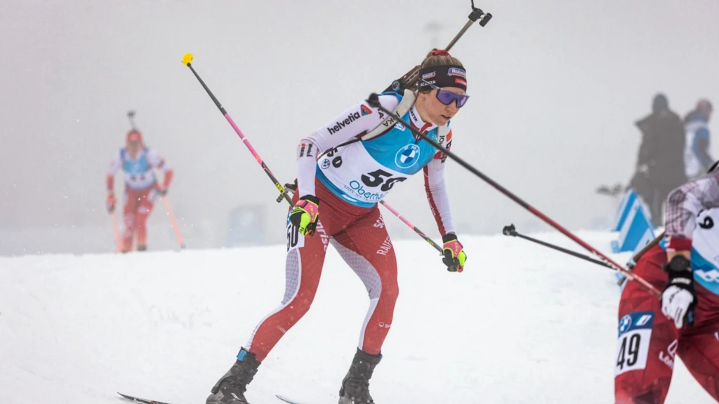 Eine Biathletin mit Startnummer 50 ist bei einem Biathlon-Wettkampf auf einer verschneiten Strecke unterwegs. Sie trägt Skier, Stöcke und ein Gewehr auf dem Rücken, während weitere Athletinnen im Hintergrund zu sehen sind.