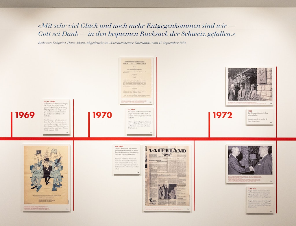Fotografie einer Museumswand mit einer rot markierten Zeitleiste der Jahre 1969 bis 1972 zur Geschichte der Beziehungen zwischen Liechtenstein und der Schweiz. An der Wand hängen Dokumente, Zeitungsseiten, Fotos und ein Zitat von Erbprinz Hans Adam.