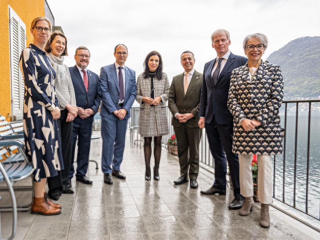 Mehrere Personen stehen anlässlich einer Vernissage nebeneinander auf einer Terrasse mit Blick auf einen See und lächeln in die Kamera. Im Hintergrund sind Berge und Wasser zu sehen.