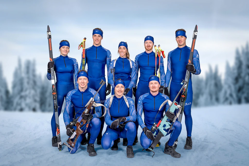 Eine Gruppe von acht Spitzensportlerinnen und Spitzensportlern des Bundesamt für Zoll und Grenzsicherheit posiert im Schnee. Einige halten Skier und Biathlongewehre, im Hintergrund sind verschneite Bäume zu sehen. Das Bild vermittelt Teamgeist und sportliche Professionalität.