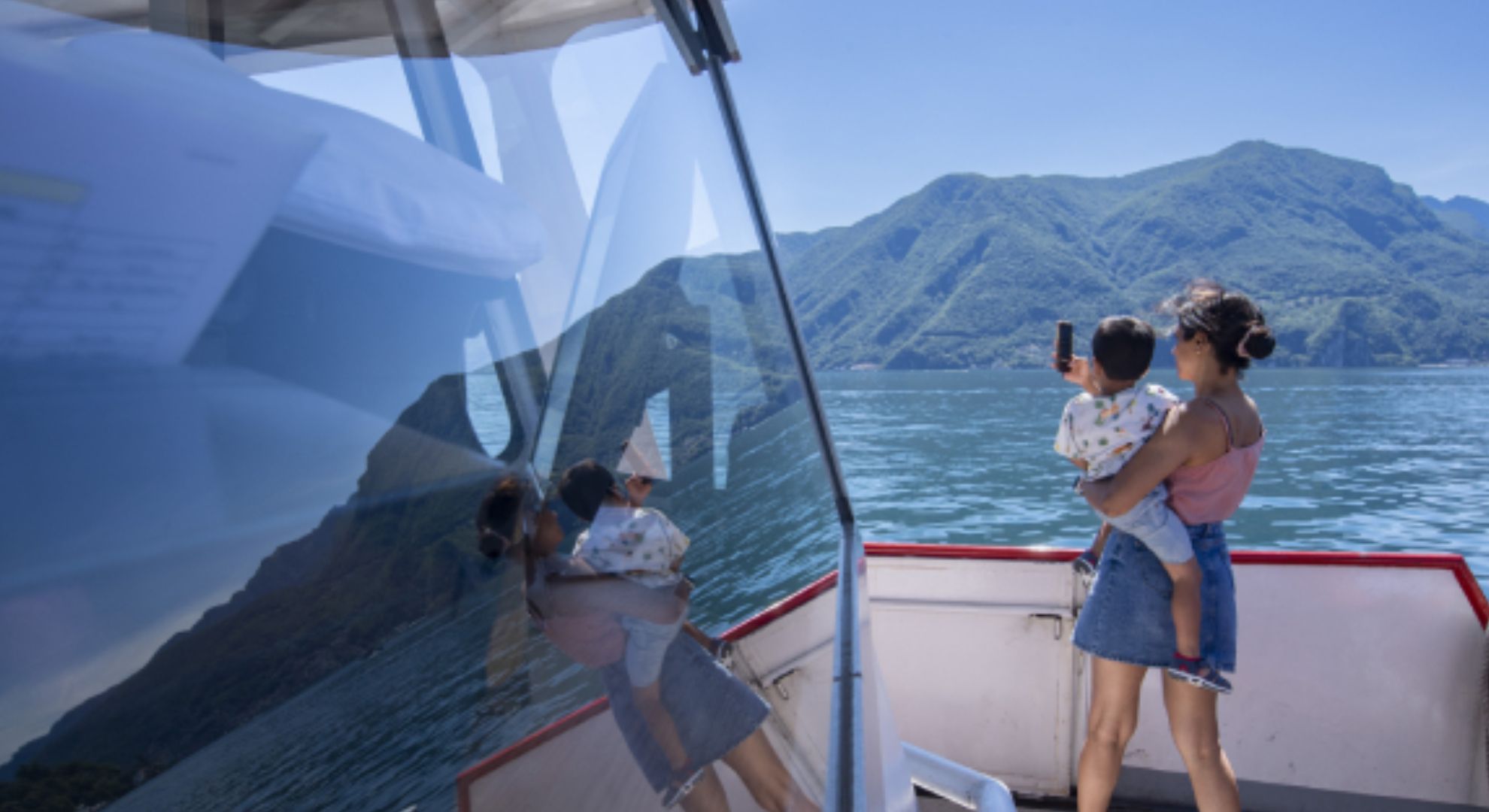 Eine Frau ist mit einem Kleinkind auf dem Schiff und macht die Überfahrt auf dem Lago di Lugano. Die Frau hält den kleinen Jungen in den Armen und macht mit ihrem Handy ein Foto des Sees. Man sieht die beiden von hinten.
