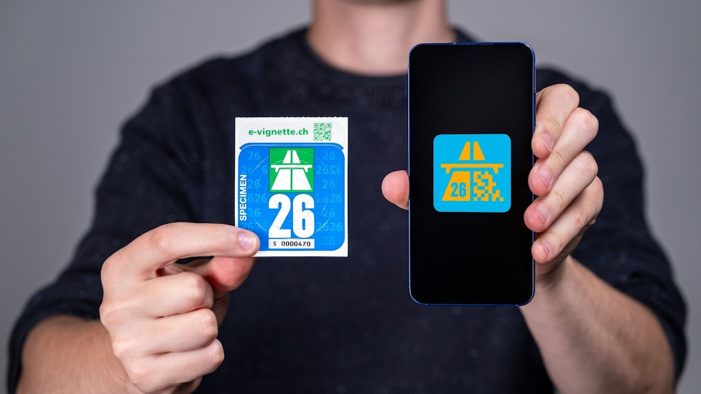 Eine Person hält eine physische Autobahnvignette und ein Smartphone mit einer digitalen Vignette. Die Vignetten sind für das Jahr 2026.