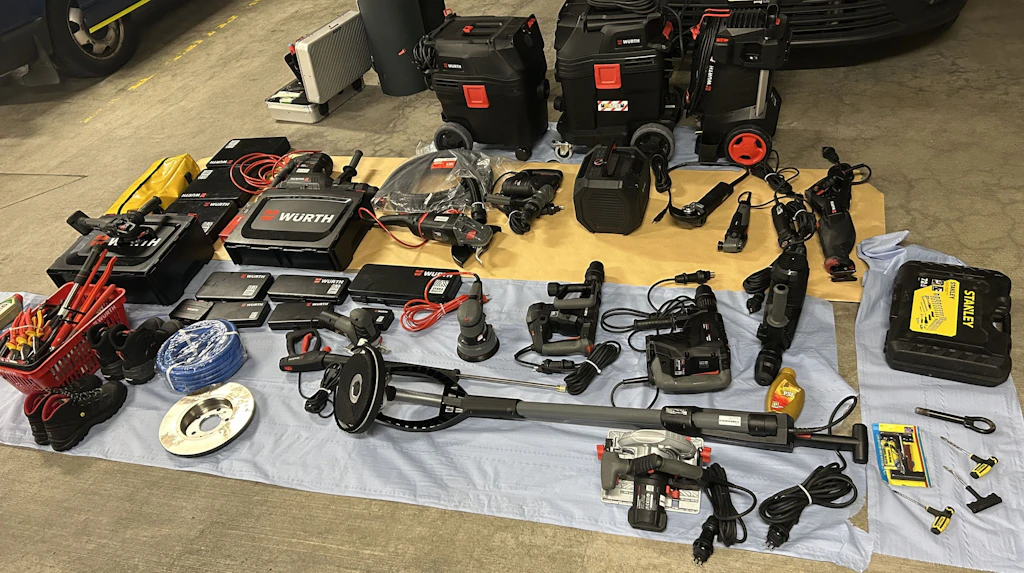 Verschiedene Elektrowerkzeuge, Handwerkzeuge und Zubehörteile sind ordentlich auf dem Boden ausgebreitet. Die Geräte stammen überwiegend von der Marke Würth und sind in Koffern, Kisten und einzeln sichtbar. Im Hintergrund sind weitere Transportboxen und ein Fahrzeug zu erkennen.