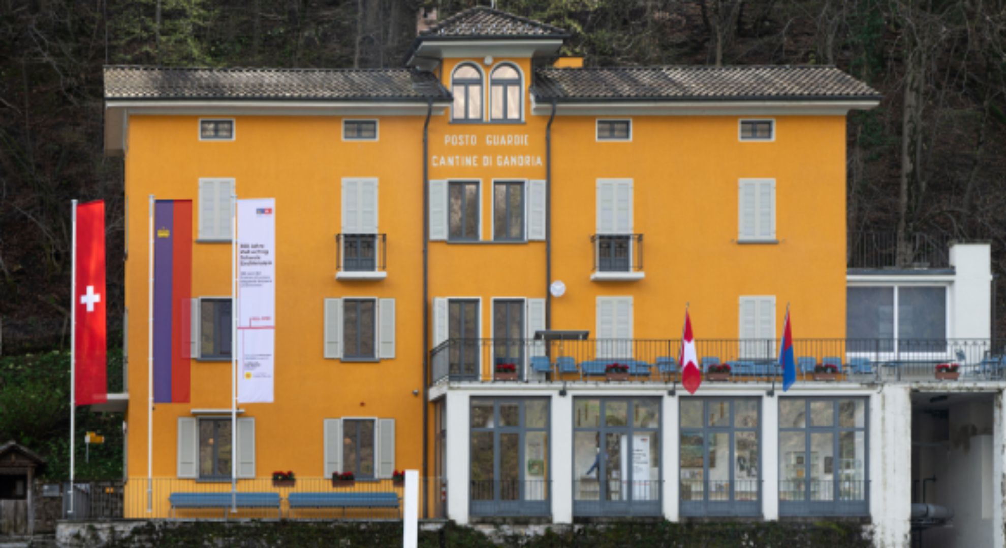 Das Bild zeigt das Zollmuseum in Gandria, ein auffälliges gelbes Gebäude mit mehreren Flaggen, darunter die Schweizer Flagge. Es ist von Bäumen umgeben und hat einen Balkon mit Sitzgelegenheiten.  