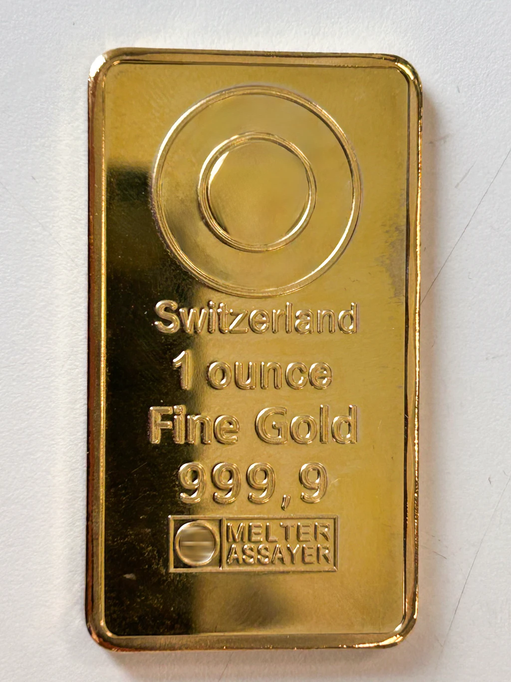 Ein rechteckiger Goldbarren mit der Prägung "Switzerland 1 ounce Fine Gold 999,9" liegt auf einer hellen Oberfläche. Die Vorderseite zeigt das Gewicht, die Feinheit und das Herkunftsland. Ein Prüfstempel ist ebenfalls sichtbar.  